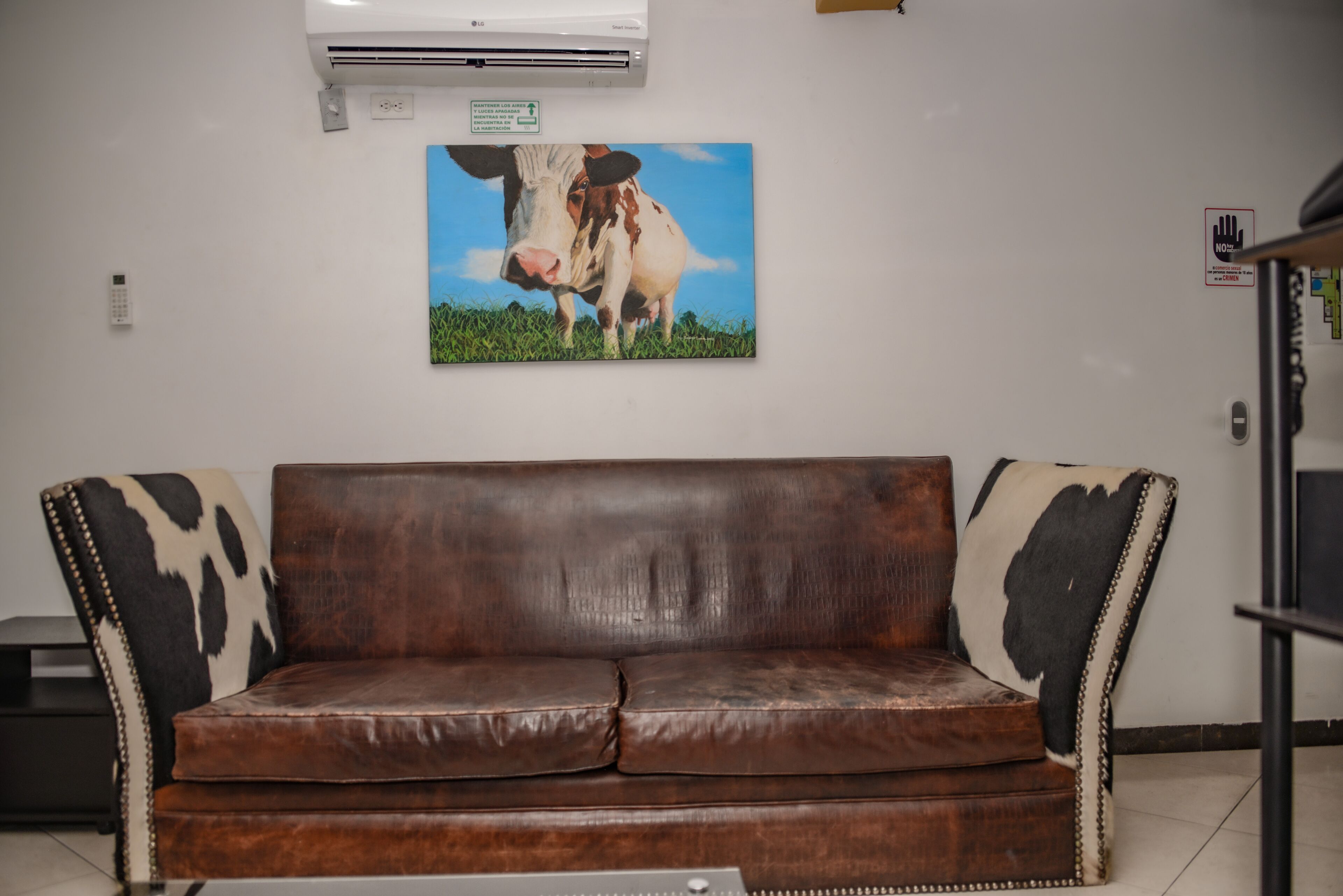Un sofá de cuero marrón con apoyabrazos de piel de vaca, un aire acondicionado instalado en la pared y un cuadro de una vaca enmarcado.