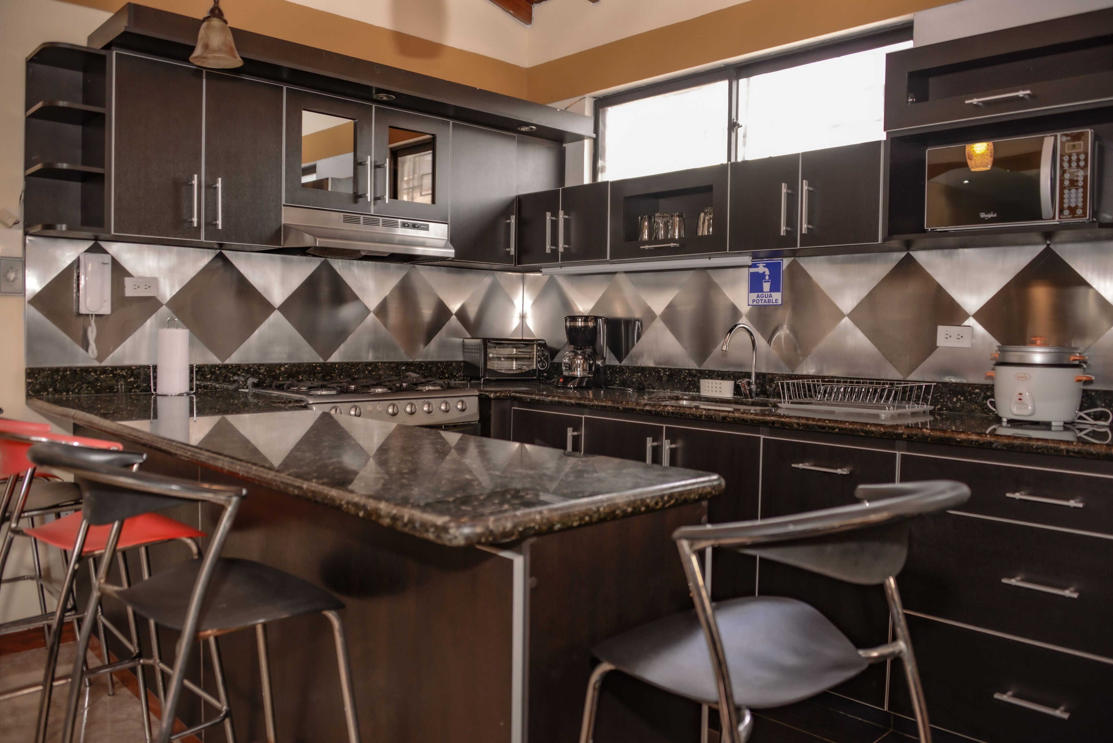 Una cocina moderna con armarios oscuros, encimera de granito y electrodomésticos de acero inoxidable. Hay dos taburetes de bar y un microondas en la encimera.