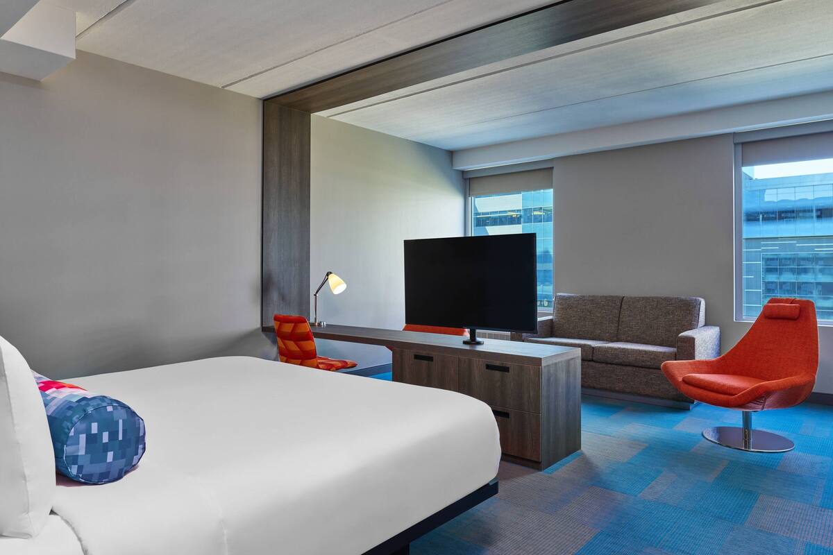 Quarto de hotel com cama, escrivaninha, televisão de tela plana, sofá e uma poltrona laranja vibrante.