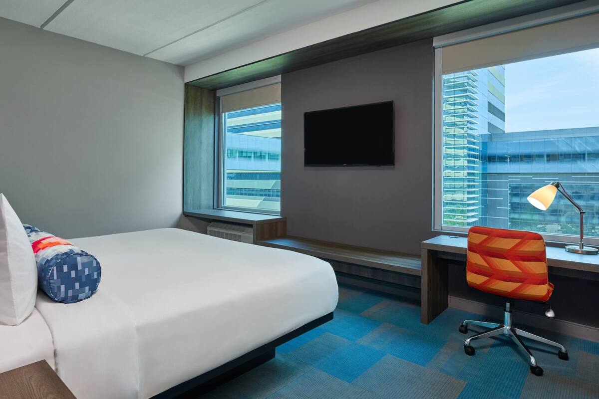 Quarto de hotel com cama, escrivaninha, cadeira, televisão e janela com vista para a cidade.