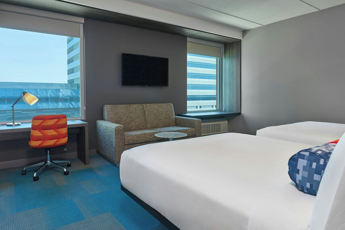 Una habitación de hotel con cama, escritorio, silla, televisión y ventana con vista a la ciudad.