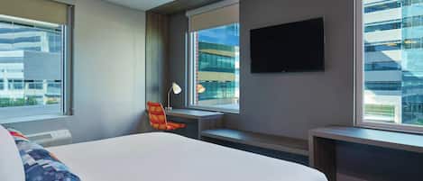 Habitación de hotel con una cama grande, un escritorio, una silla, un televisor y una ventana con vistas a la ciudad.