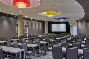 Uma sala de conferências com fileiras de mesas e cadeiras, uma tela grande e um palco.