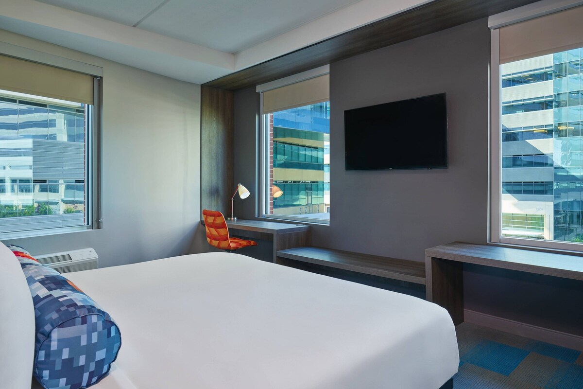 A hotel room with a large bed, a desk, a chair, a TV, and a window with a city view.
