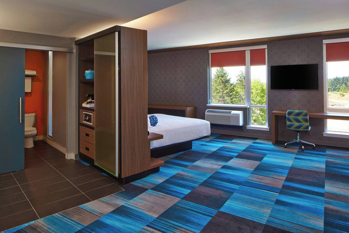 A hotel room with a bed, a desk, a chair, a TV, and a window with a view of trees.