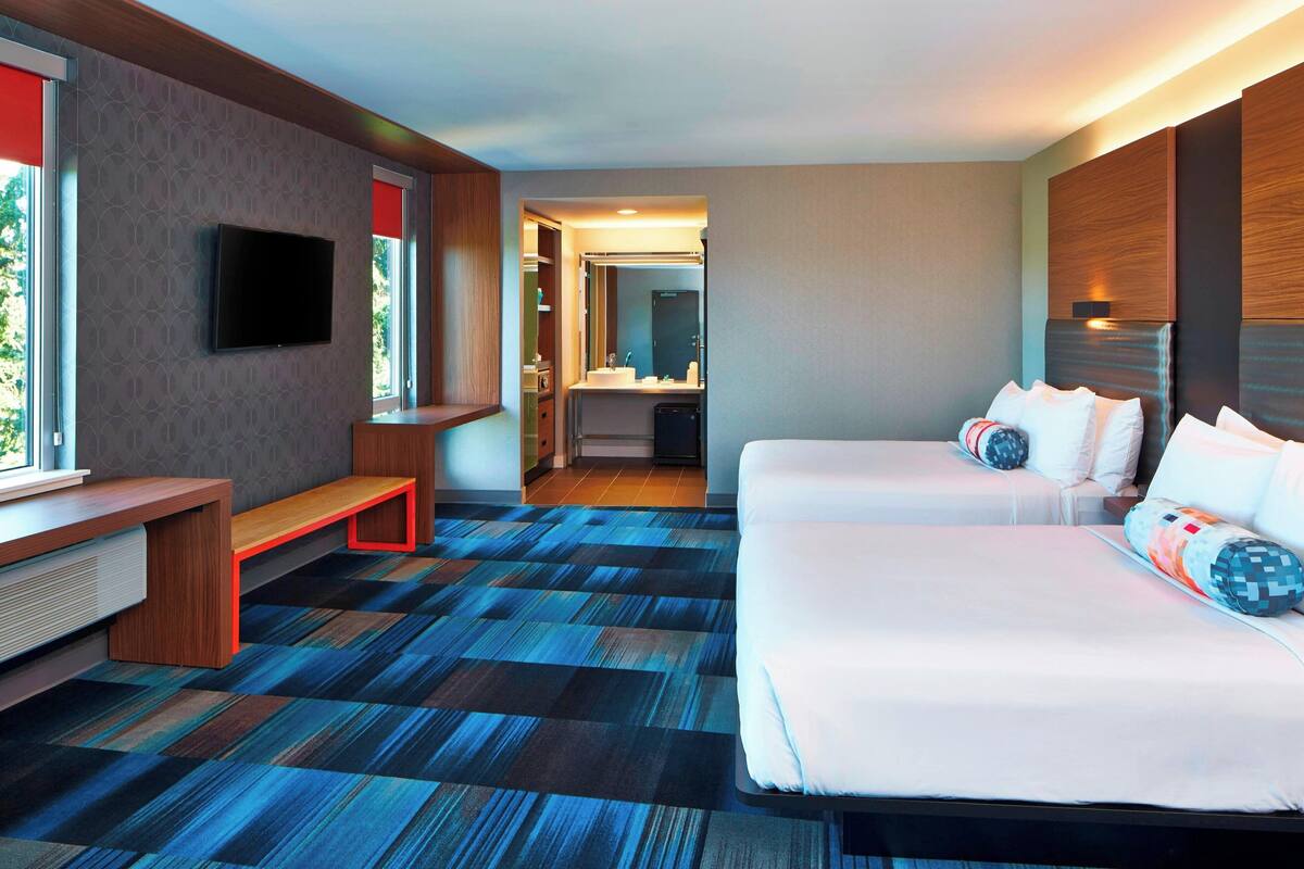 A hotel room with a large bed, a bench, a TV, and a bathroom visible through an open door.
