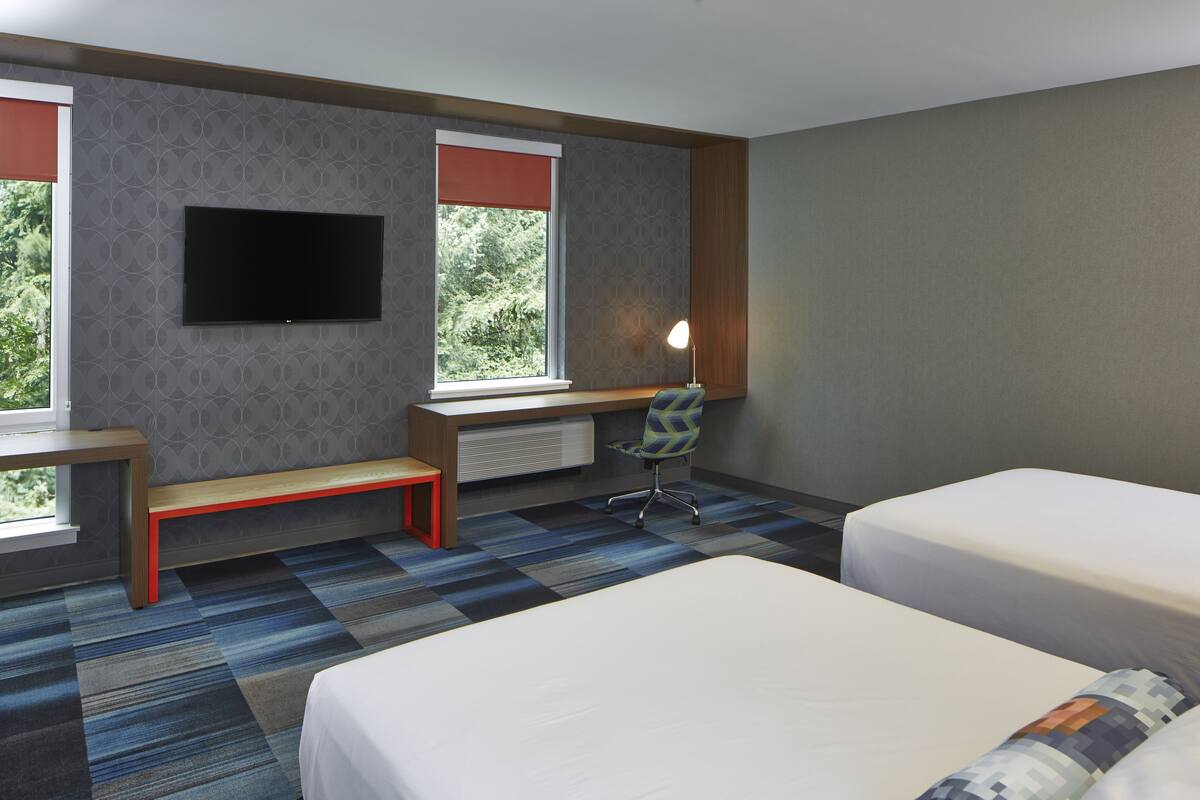A hotel room with two beds, a desk, a chair, a TV, and a window with a view of trees.