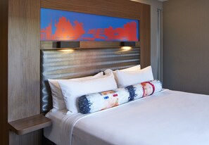 A neatly made bed with white linens and a colorful pillow, a wooden headboard with a cityscape artwork, and a small wooden nightstand.