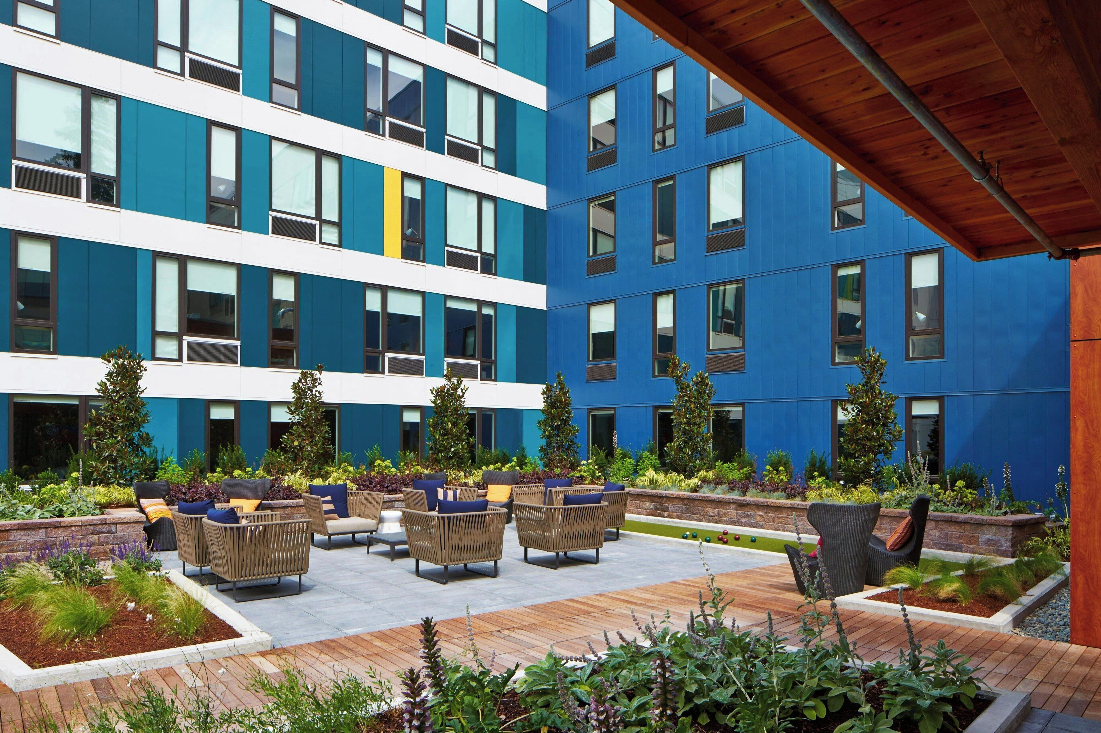 Un edificio moderno con paredes exteriores azules y blancas, un área de asientos al aire libre con muebles de mimbre y jardines paisajísticos.