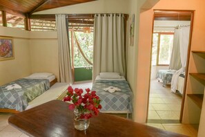 Ein Zimmer mit zwei Betten, einem Holztisch mit Blumenvase und Blick auf einen Balkon mit Vorhängen.