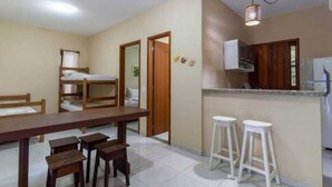 Habitación pequeña con una litera, una mesa de comedor con sillas, una zona de cocina con mostrador y taburetes, y una lámpara colgante.