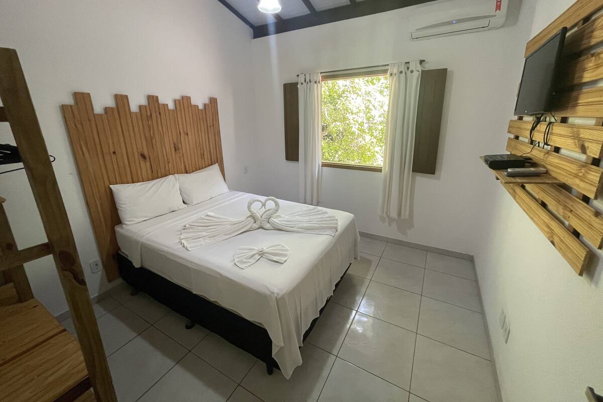Habitación de hotel con cabecera de madera, cama con ropa blanca, ventana con cortinas, televisor de pantalla plana montado en un panel de madera y piso de baldosas.