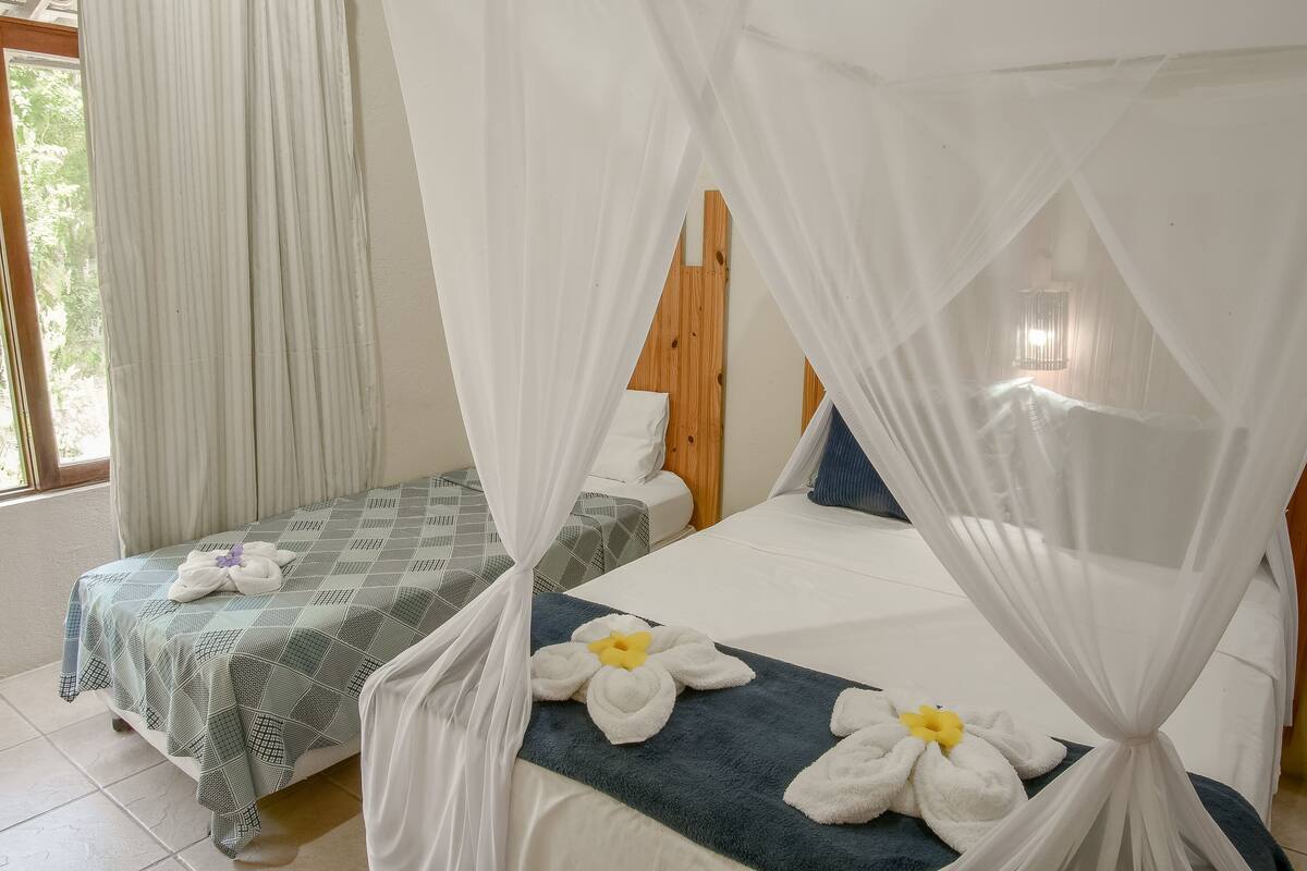Habitación de hotel con dos camas, cada una con dosel, y un ventanal con cortinas.