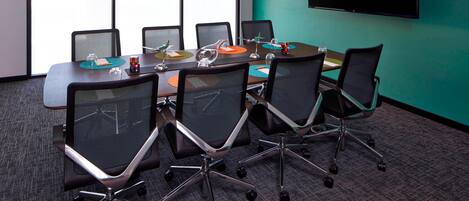 Un salón de conferencias con una mesa grande, cuatro sillas de oficina, un televisor de pantalla plana y un amplio ventanal con persianas.