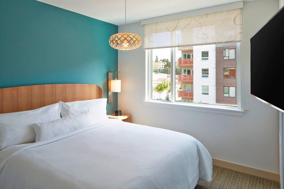 Habitación de hotel con una cama grande, cabecera de madera, lámpara de noche, ventana con vista a edificios y televisión en la pared.