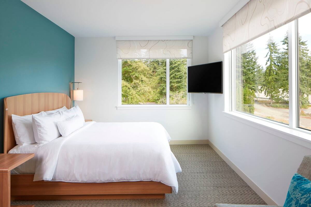 Un dormitorio con una cama, una mesita de noche, un televisor montado en la pared y una ventana con vistas a árboles.