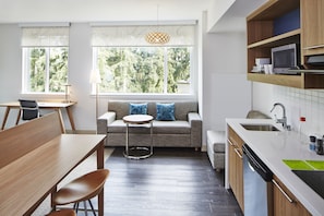 A modern kitchen with a dining area, a sofa with blue cushions, and a view of trees through large windows.