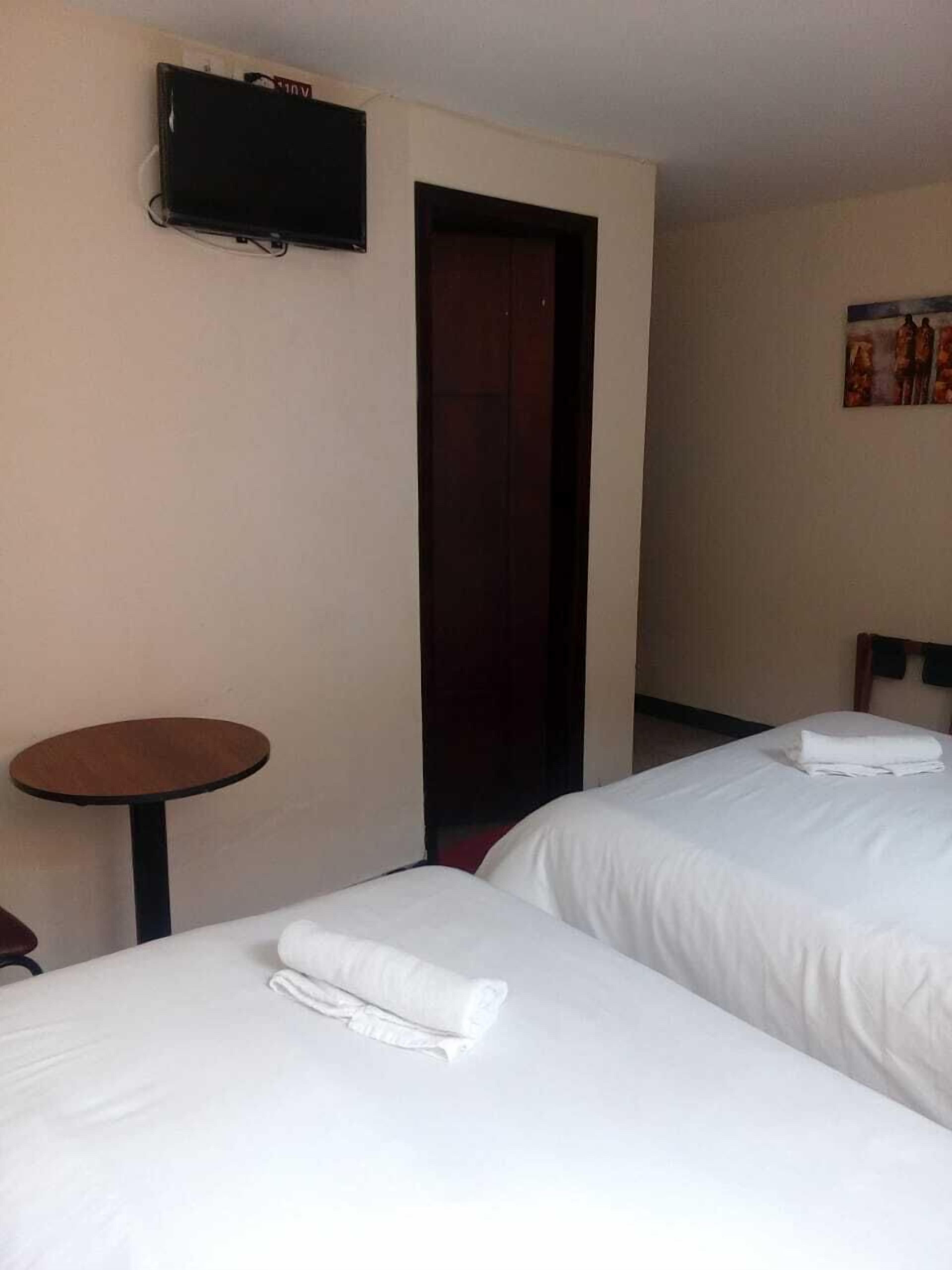 A hotel room with two beds, a small round table, a TV mounted on the wall, and a door leading to another room.