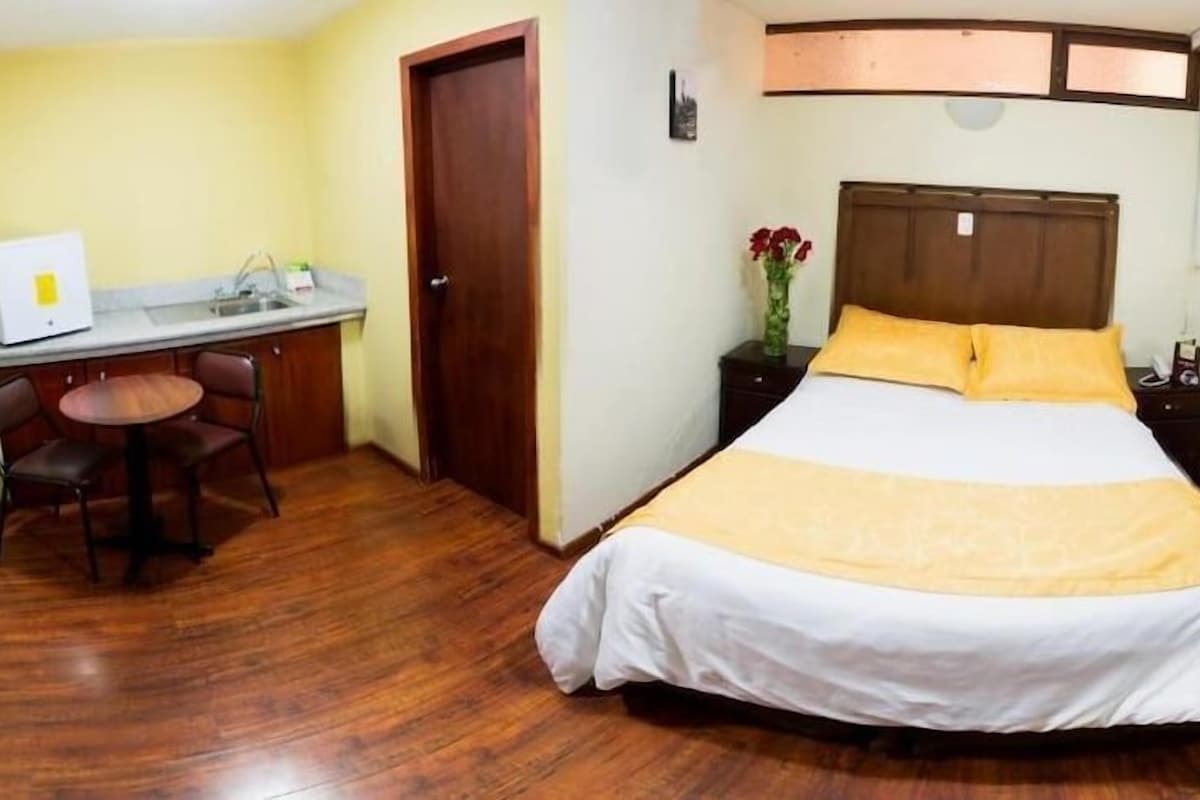 Habitación de hotel con cama, escritorio, silla, mesita de noche y un jarrón con flores.