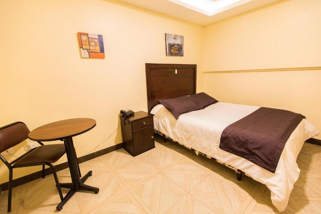 A small, neatly arranged hotel room with a bed, a bedside table, a chair, and a small table.