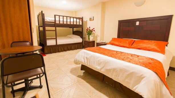 Habitación de hotel con literas, una cama individual con ropa de cama naranja, una mesita pequeña con una silla y una mesita de noche con un jarrón de flores.
