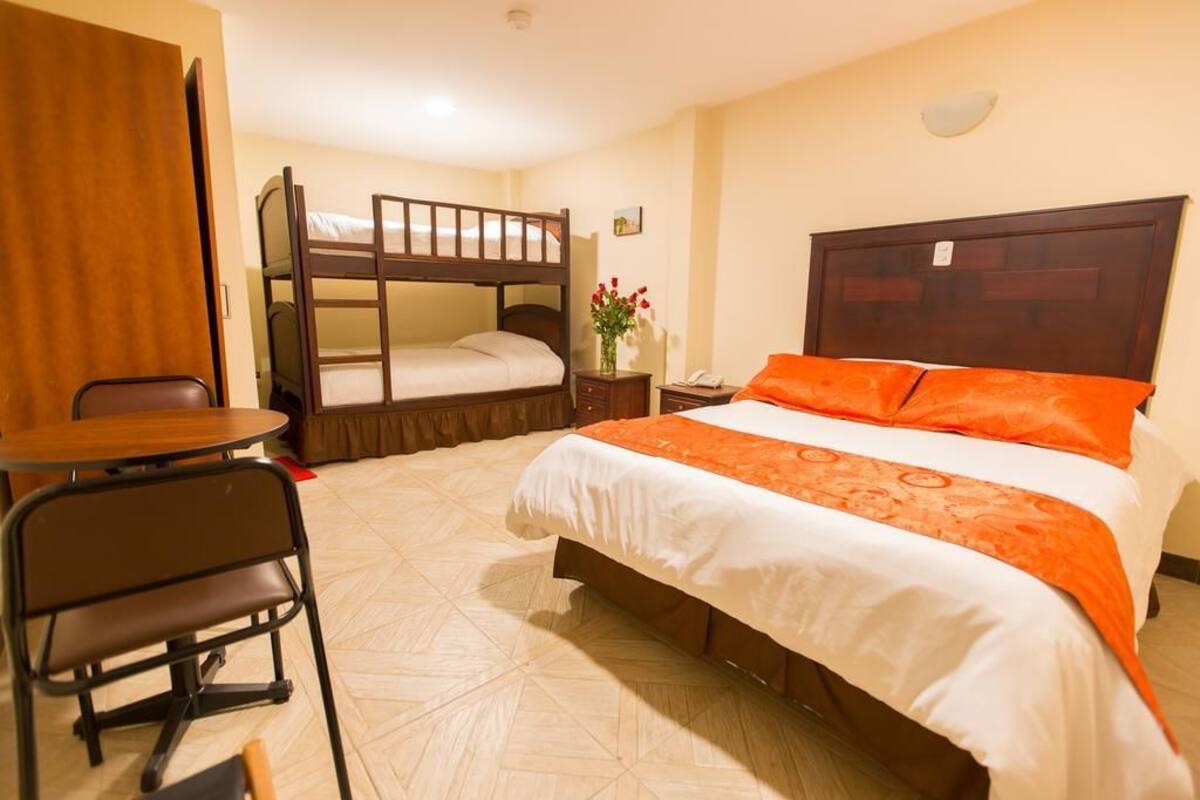 Habitación de hotel con literas, una cama individual con ropa de cama naranja, una mesita pequeña con una silla y una mesita de noche con un jarrón de flores.