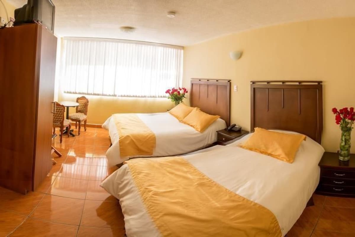Habitación de hotel con dos camas, un armario de madera, una mesita con sillas y una ventana con cortinas.