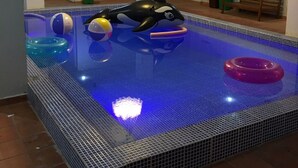 Una piscina interior pequeña con un flotador con forma de tiburón, un banco y un niño jugando.