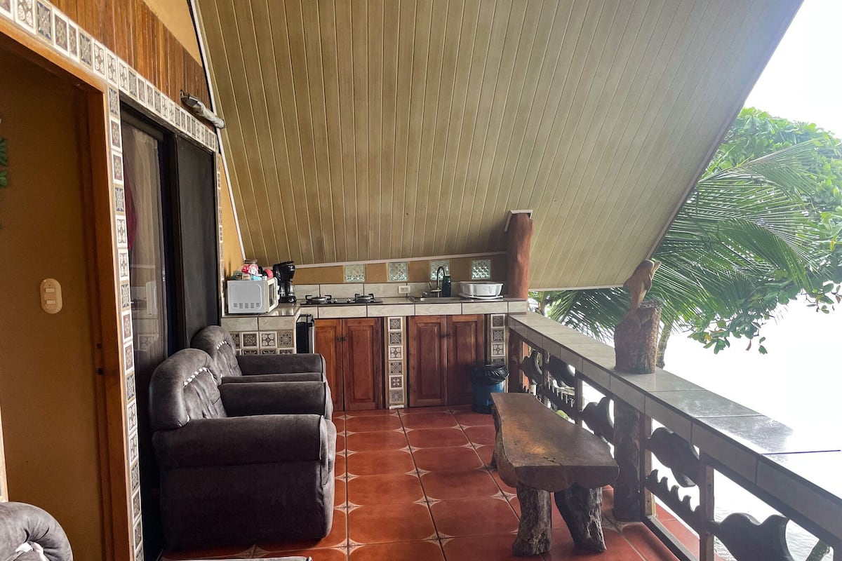 A kitchen with a tiled backsplash, wooden cabinets, and a countertop with various appliances. There is a dining area with a bench and a table, and a balcony with a railing overlooking a body of water.