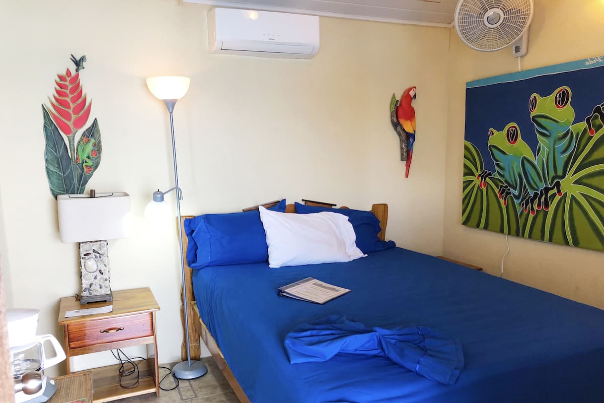 A neatly made bed with blue bedding, a standing lamp, a wall-mounted air conditioner, a fan, and a colorful painting of frogs.