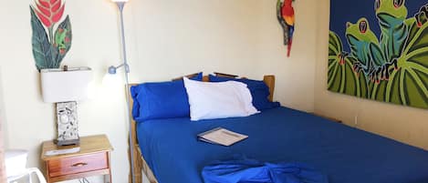 Una cama bien tendida con ropa de cama azul, una lámpara de pie, un aire acondicionado de pared, un ventilador y un colorido cuadro de ranas.