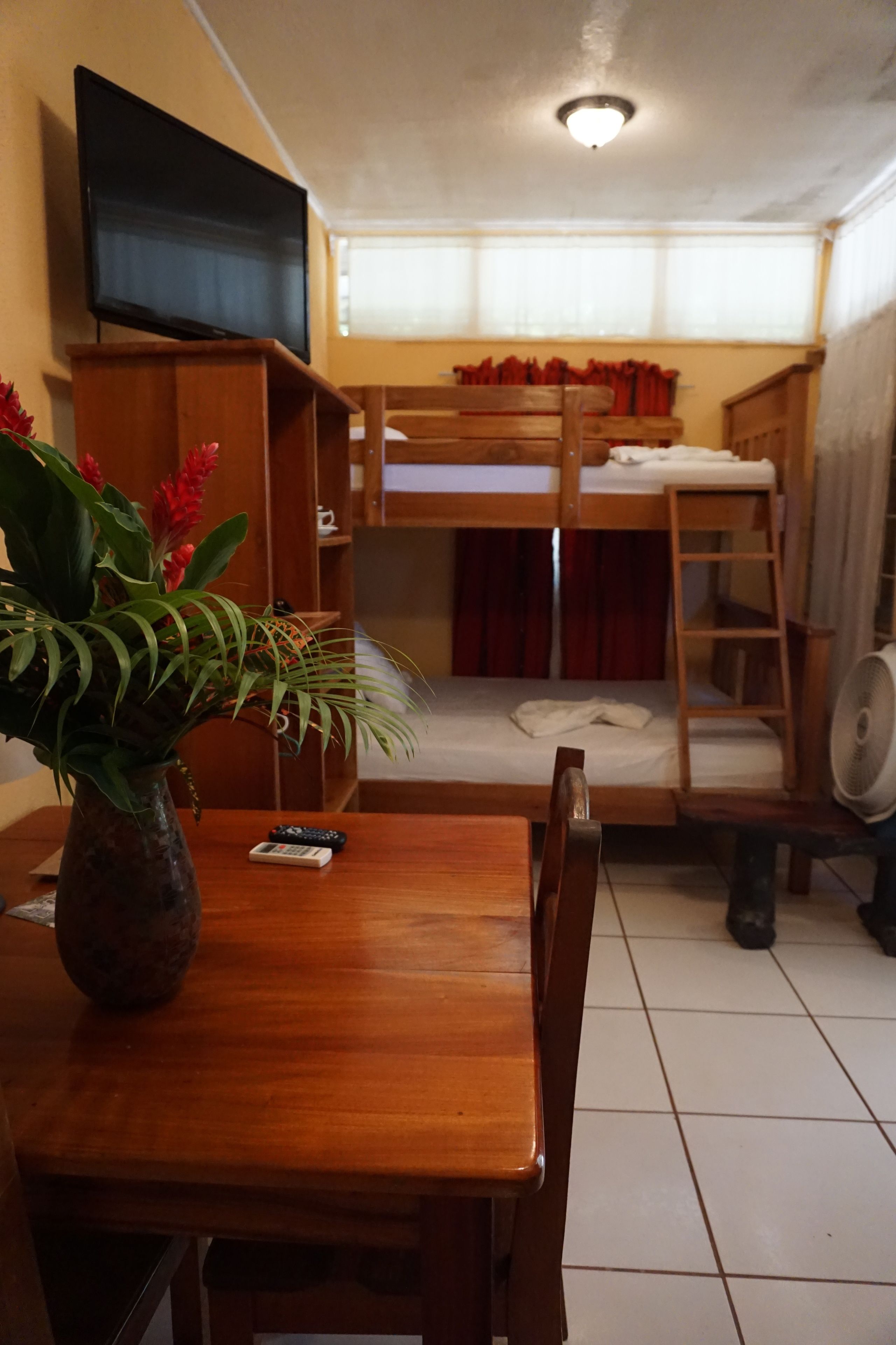 Una habitación con una cama litera de madera, un televisor montado en la pared, una mesa de madera con un florero y un ventilador en el suelo.