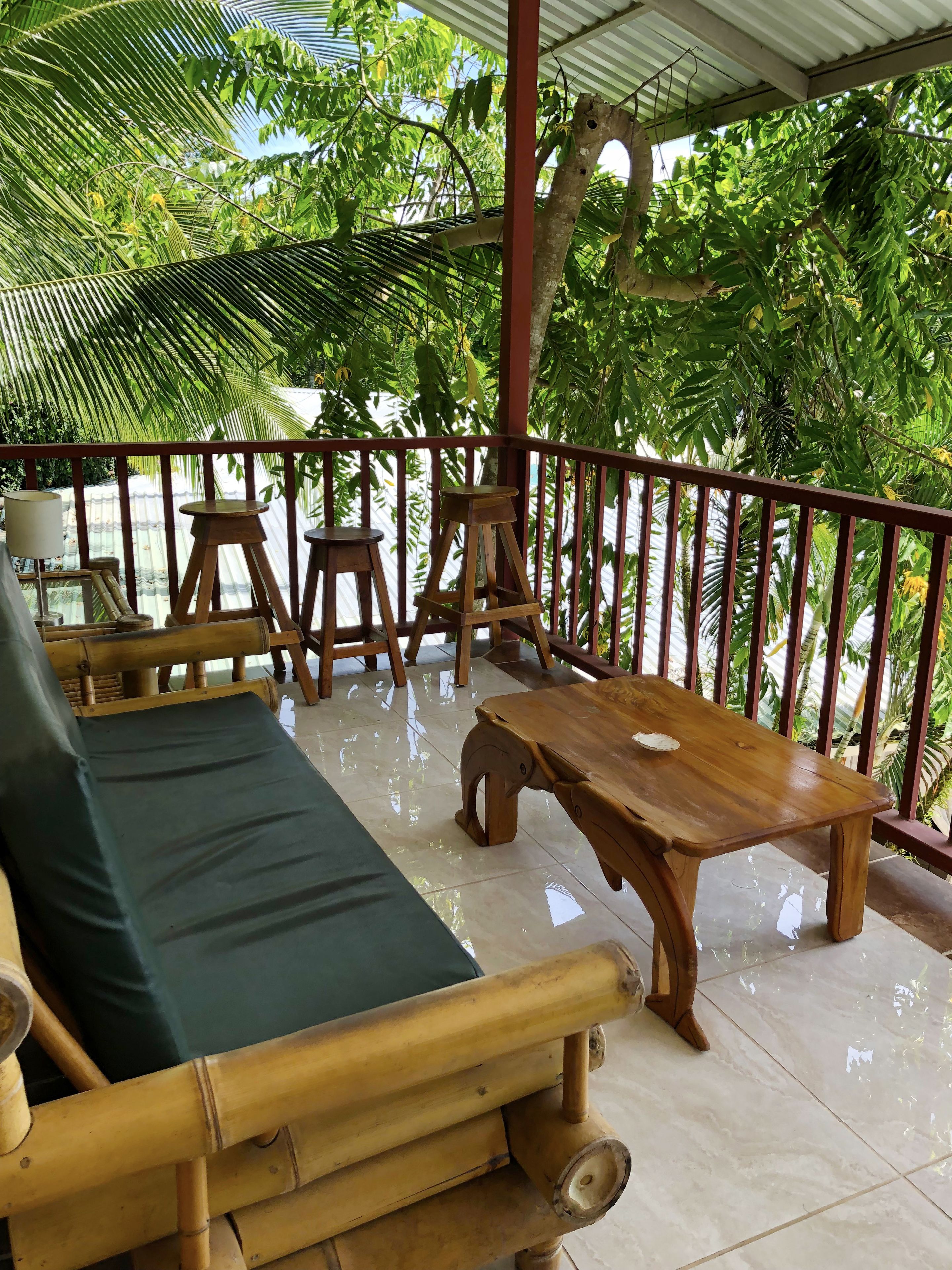 A wooden lounge chair and table set on a tiled patio with a metal railing, surrounded by lush greenery.
