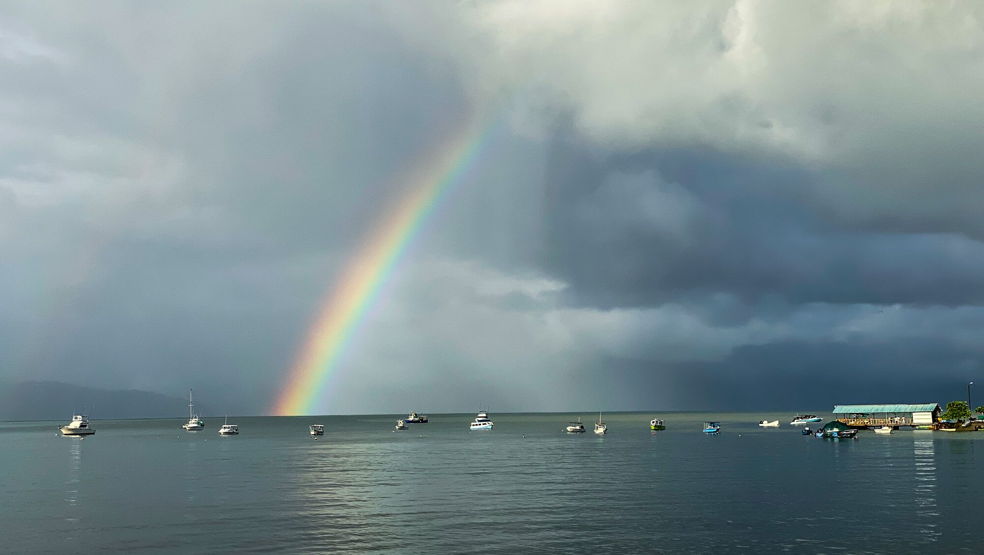 A coastal scene with a rainbow, boats, and a building with a green roof.