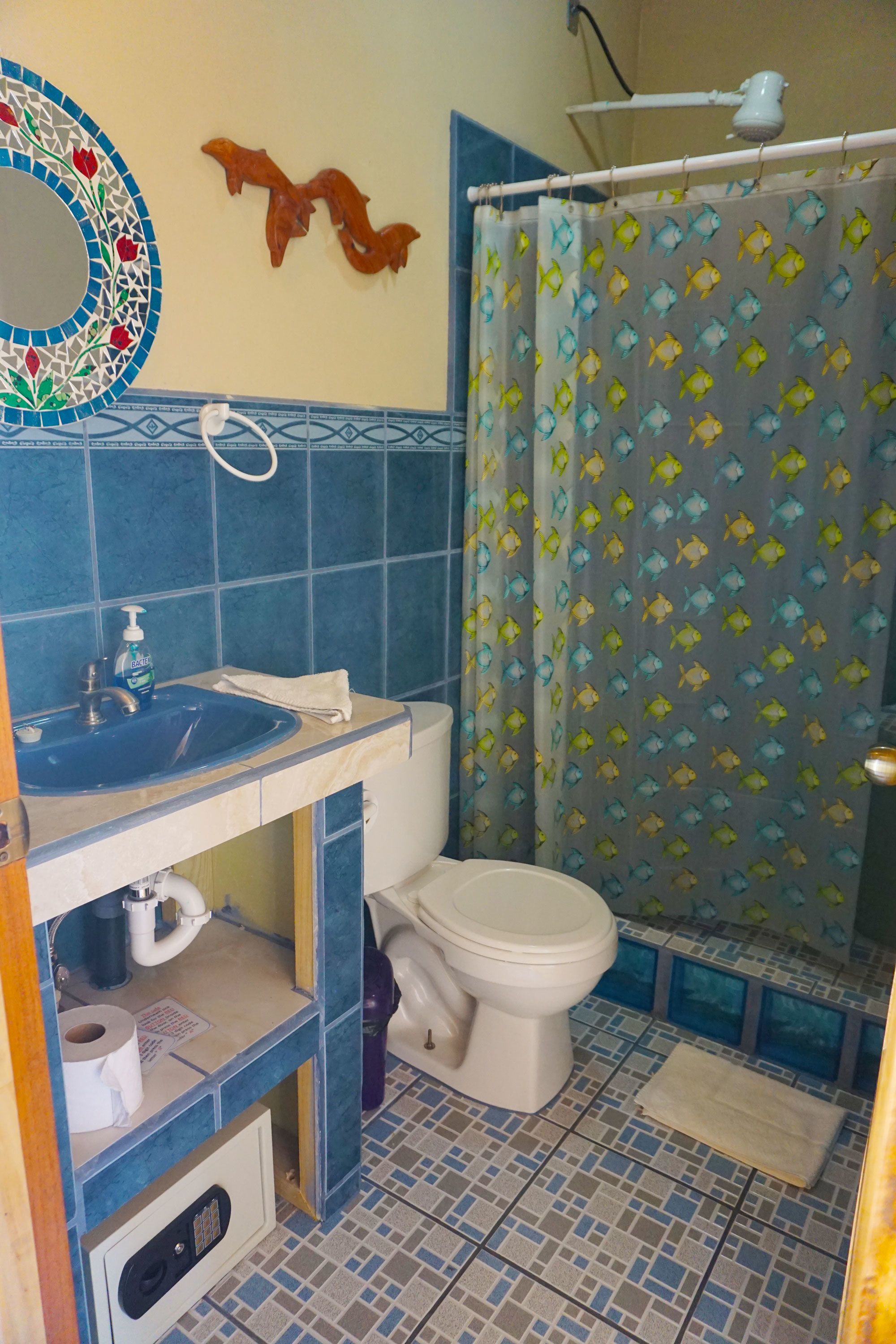 Un baño con piso de azulejos azules, un inodoro blanco, un lavamanos azul y una cortina de ducha con estampado de peces.