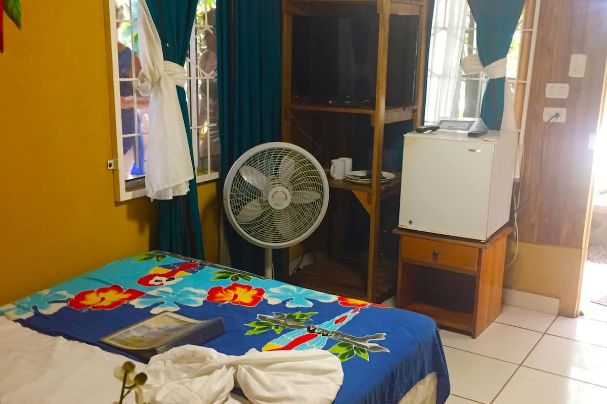 A hotel room with a bed, a fan, a wooden cabinet, and a window with curtains.