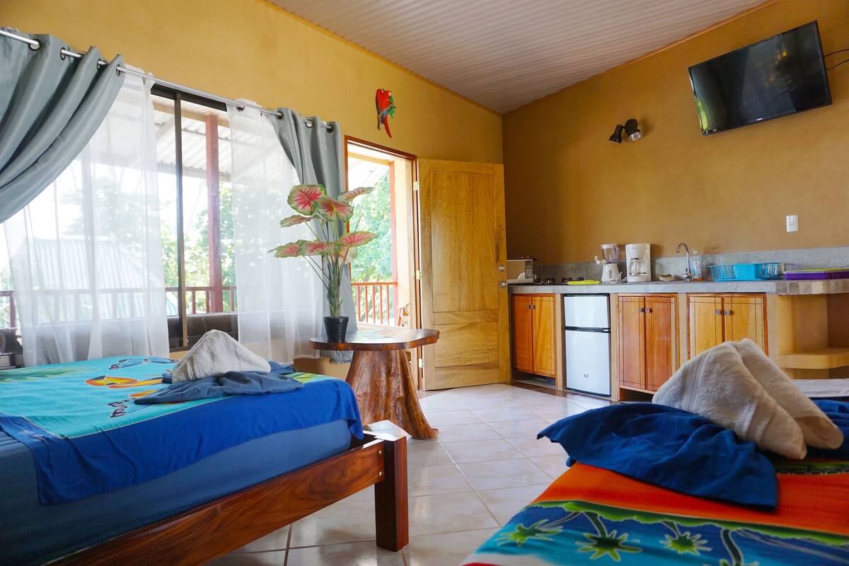 A room with two beds, a small kitchen area, and a window with curtains.