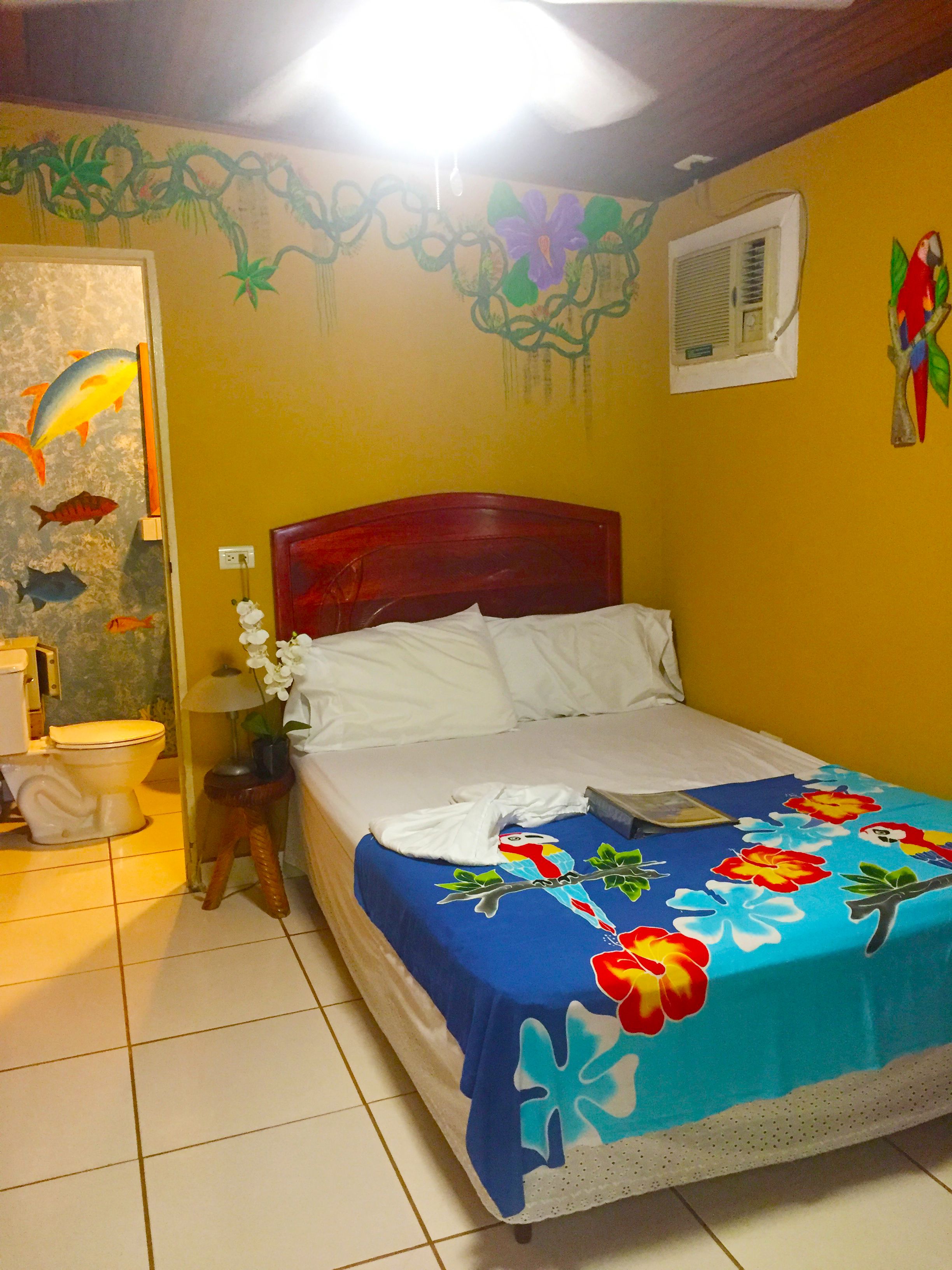A bedroom with a wooden headboard, a bed with a blue floral bedspread, a white pillow, and a nightstand with a lamp.