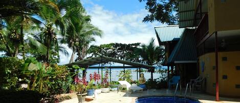 A pool area with lounge chairs, a covered seating area, and a view of the sea.