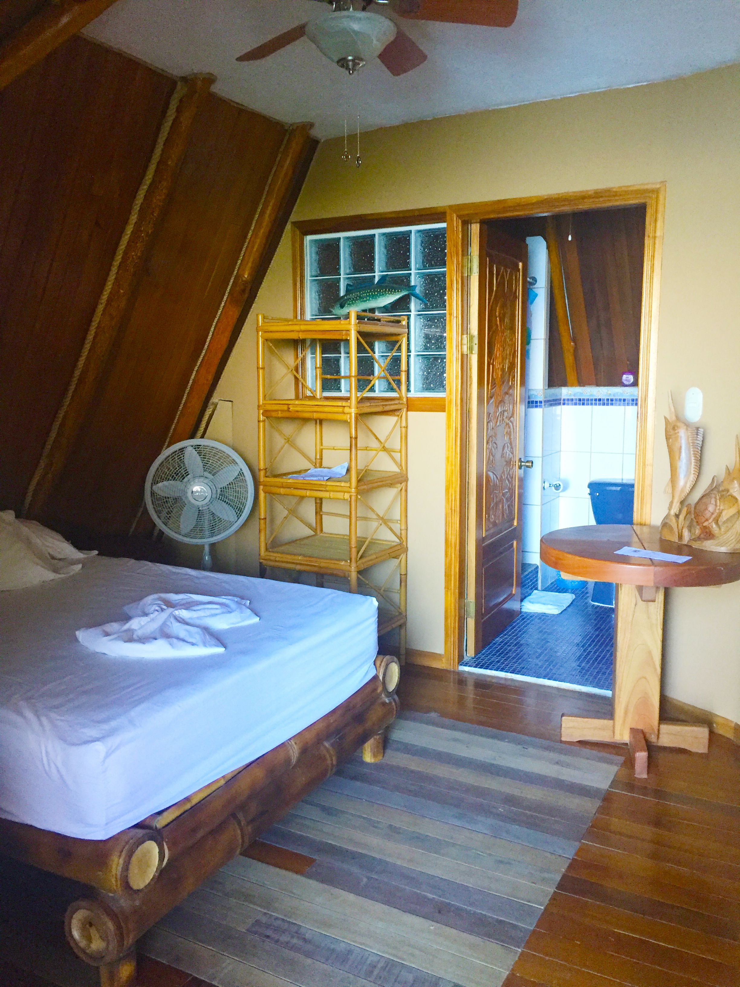 A bedroom with a bed, a fan, a wooden shelf, and a door leading to a bathroom.