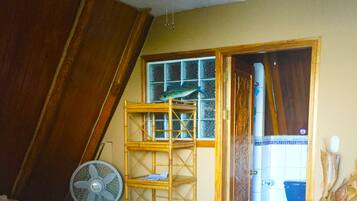 A bedroom with a bed, a fan, a wooden shelf, and a door leading to a bathroom.
