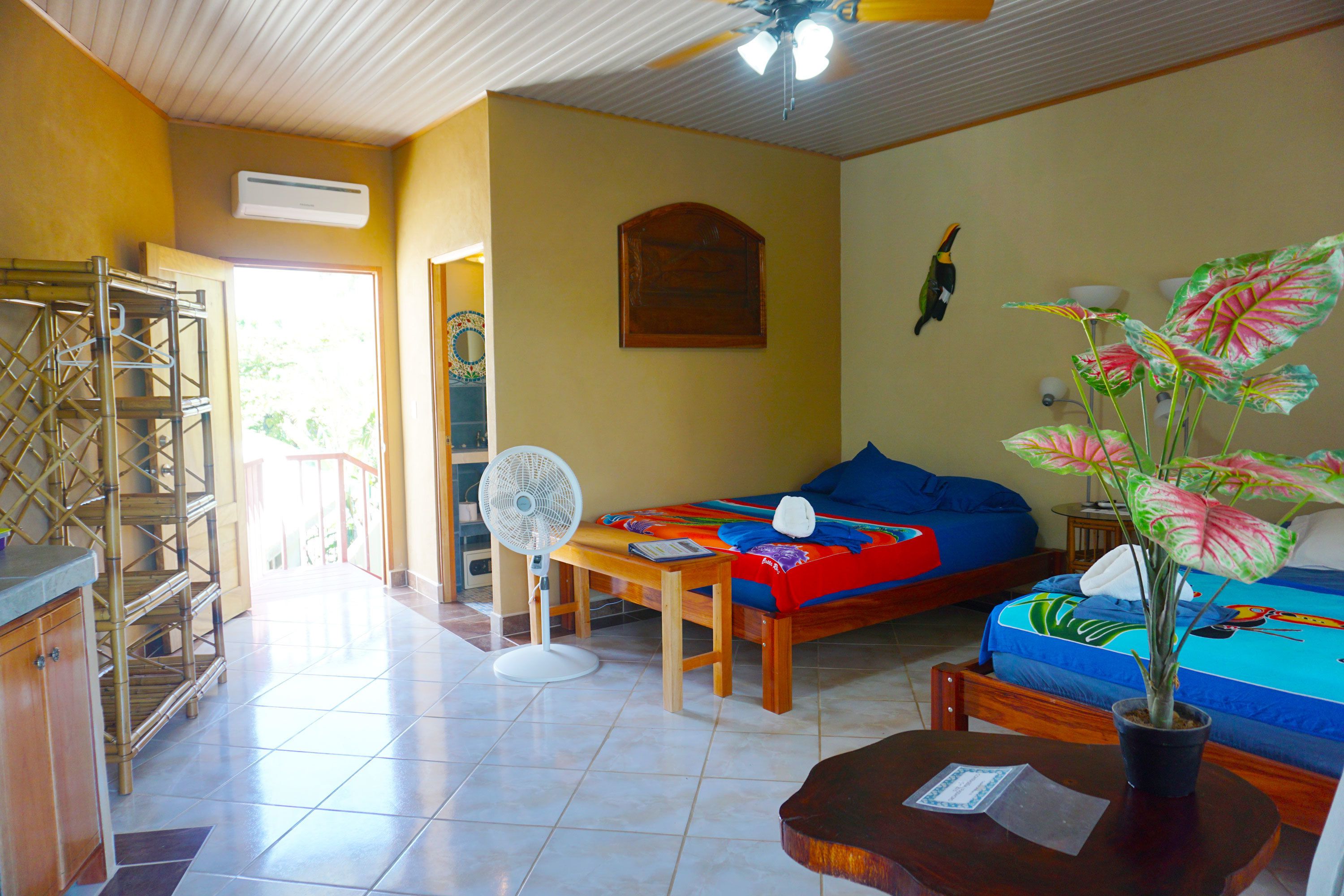 Una habitación con una cama, un ventilador, una mesa de madera, una planta en maceta y una ventana.