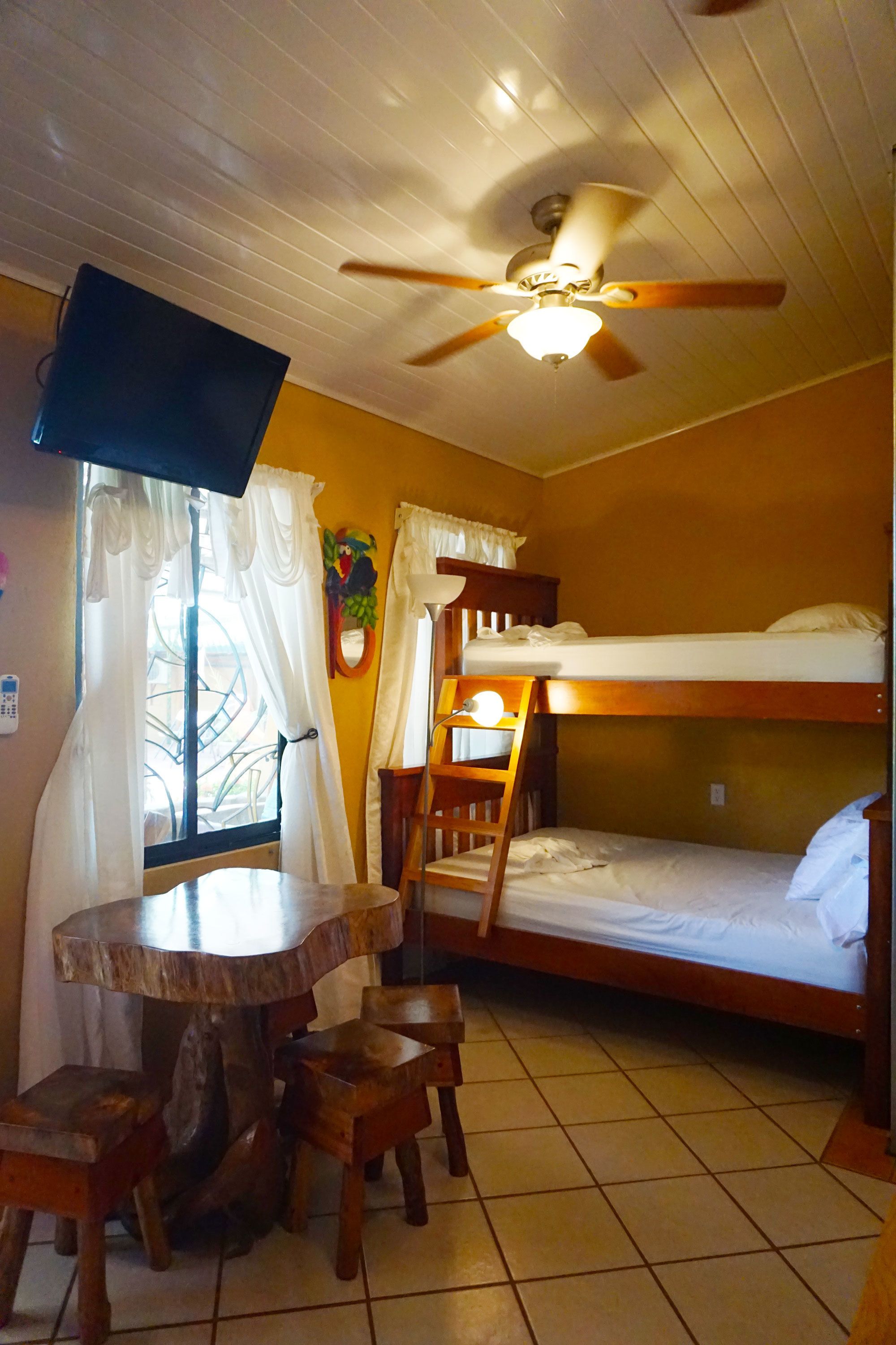 A room with bunk beds, a round wooden table with chairs, a ceiling fan, and a TV mounted on the wall.