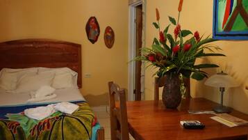 Habitación de hotel con una cama, una mesa de madera, un jarrón con flores, una lámpara y un cuadro en la pared.