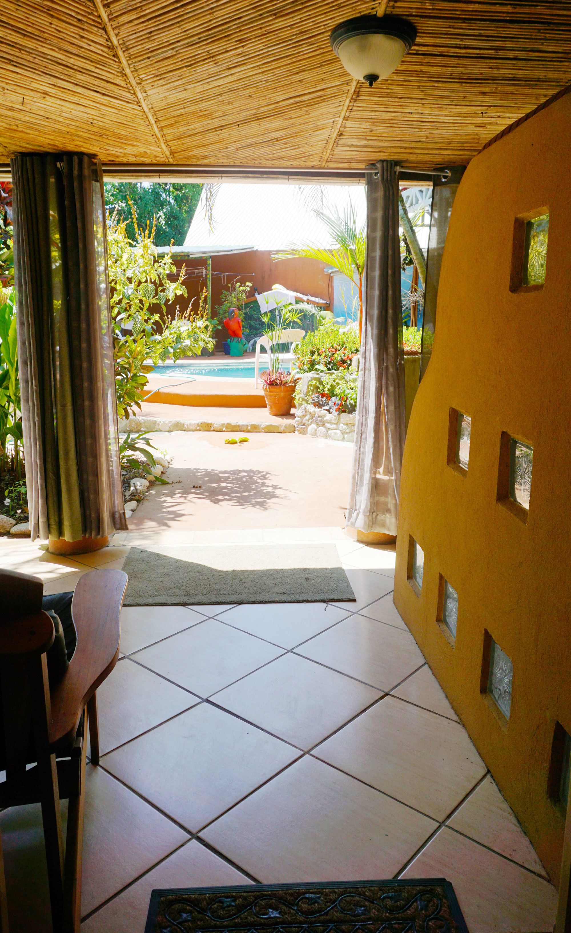 Ein Zimmer mit Fliesenboden, einem Sessel, einem Tisch, einem Deckenventilator und Blick auf einen Außenbereich mit Pool und Pflanzen.