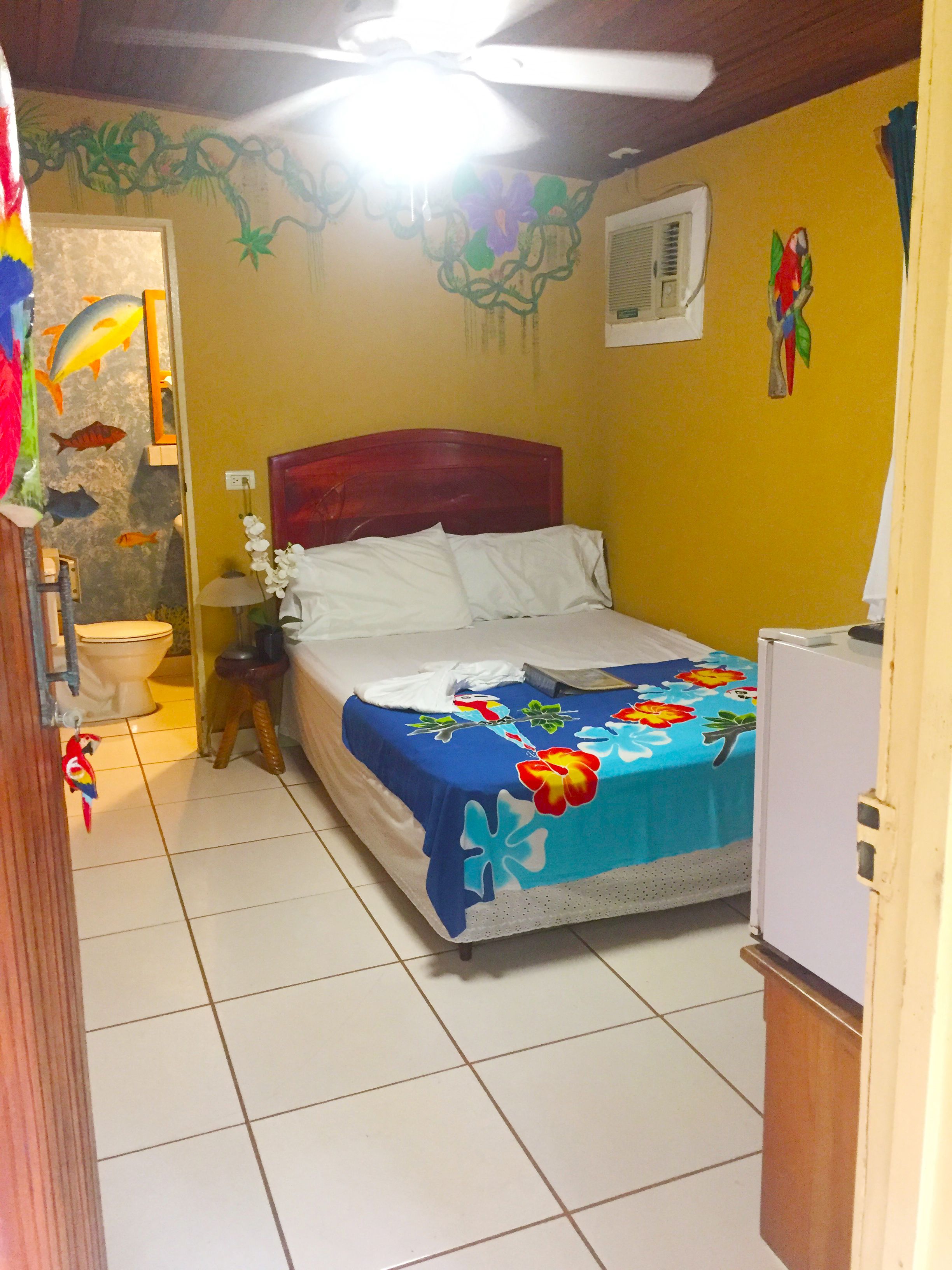 A bedroom with a bed, a nightstand, a lamp, a toilet, and a refrigerator.