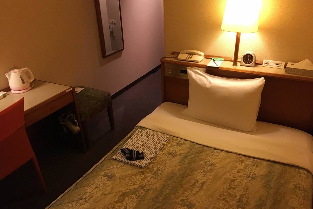 Een hotelkamer met een bed, een bureau met telefoon, een lamp en een wekker op het nachtkastje.