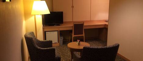 A hotel room with two patterned armchairs, a small round table, a television on a stand, and a desk with a chair.