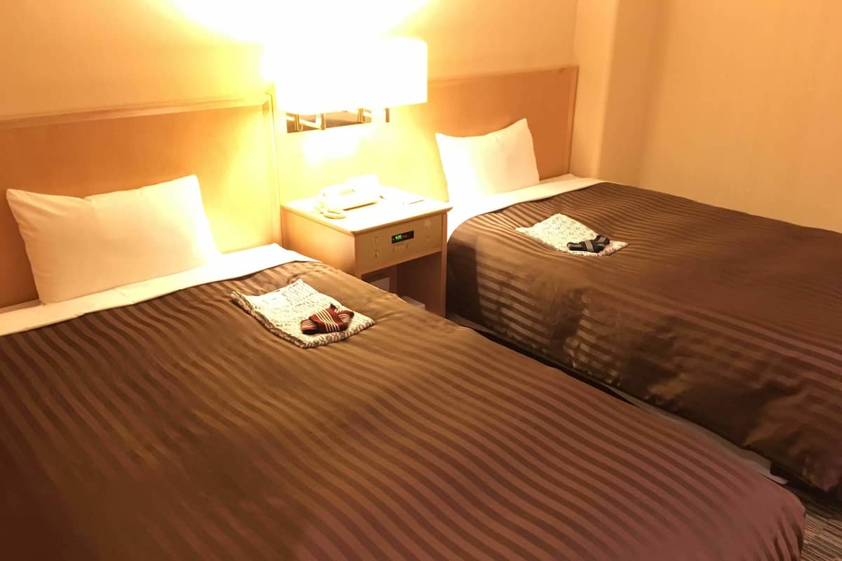 A hotel room with two beds, each with a pillow and a folded towel. There is a nightstand with a lamp between the beds.
