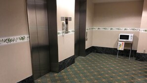 A hotel corridor with a patterned carpet, a microwave on a stand, and an elevator with a phone. - Kashiwa Plaza Hotel Annex (Kashiwa)